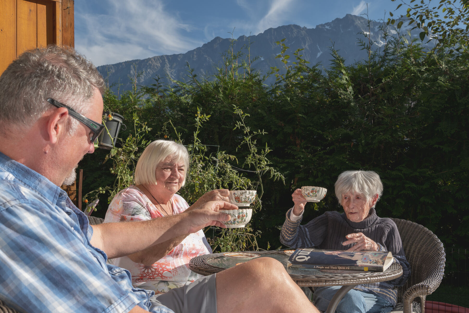 An Evergreen provider sharing tea with two clients on a sunny patio, mountains behind